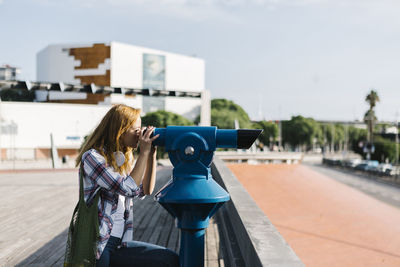 Young woman looking through binoculars while sitting on footpath during sunny day