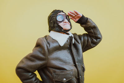 Senior woman wearing glasses while saluting against yellow background