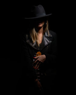 Female model wearing hat while holding guitar against black background