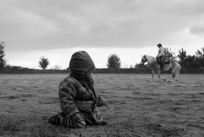 Baby boy looking at woman riding horse on field against sky