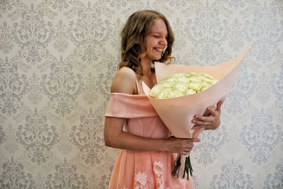 Smiling woman holding bouquet while standing against wall