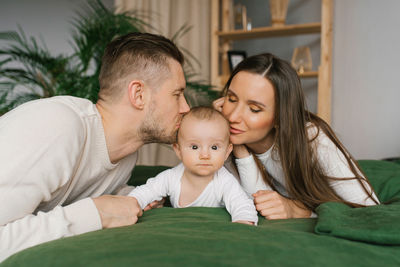 Young happy parents kiss their baby son lying on the bed