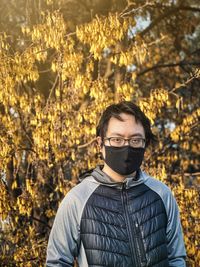 Portrait of young asian man in eyeglasses and face mask standing against flowering laburnum tree.