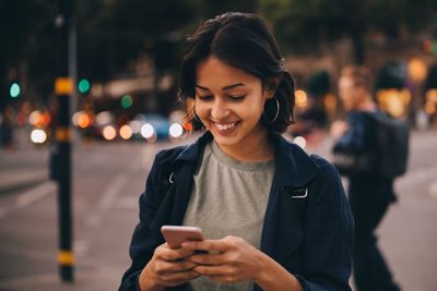 Smiling young woman using social media on phone while standing in city