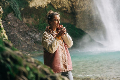 A young woman drinking a hot drink in a mountain canyon in a nature reserve near a large waterfall.