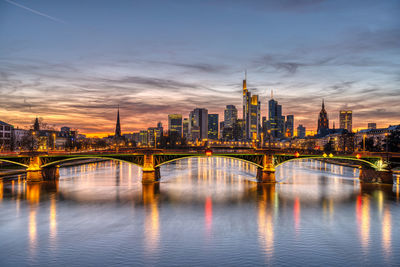 Bridge over river by buildings against sky at dusk