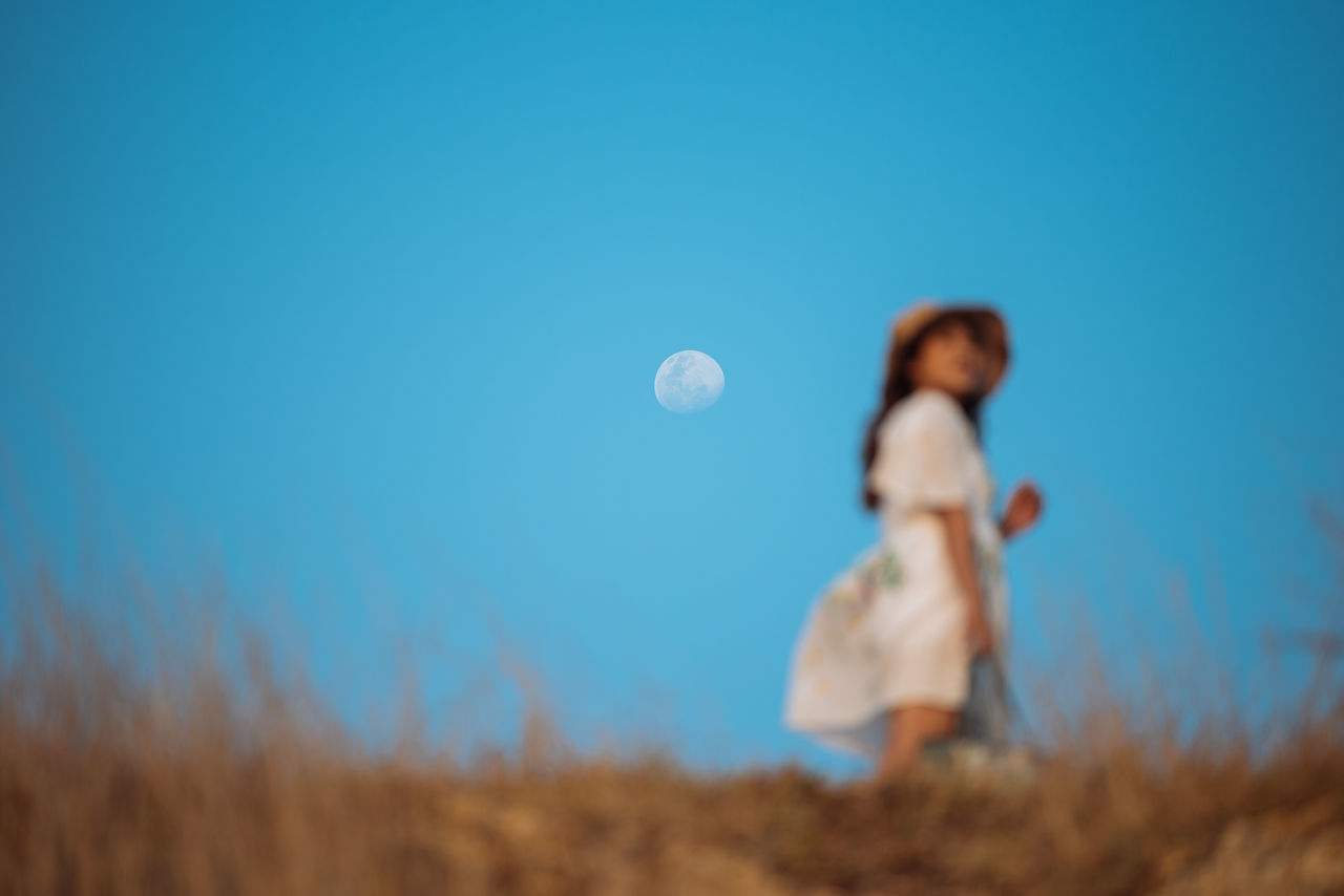 sky, land, nature, moon, plant, field, one person, blue, leisure activity, selective focus, standing, grass, real people, full length, young adult, women, lifestyles, copy space, clear sky, outdoors