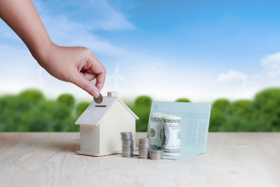 Close-up of hand putting coin in model house at table against sky