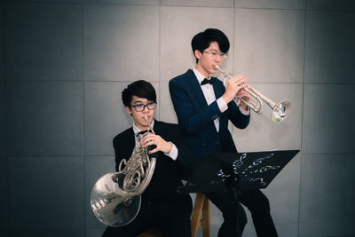 Two men playing trumpet and french horn against wall