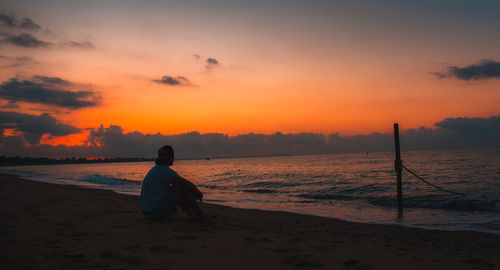 Silhouette man sitting at beach against sky during sunset
