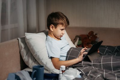 Little boy using tablet while laying in bed in living room. medicines and thermometer in foreground