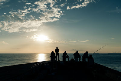 Silhouette people fishing at sea against sky during sunset