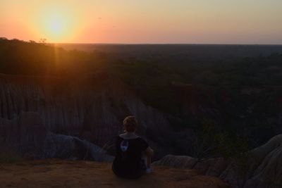 Rear view of woman sitting on rock against sky during sunset