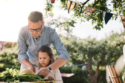 Grandfather teaching granddaughter to cut vegetable at table in backyard