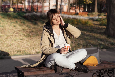 Young woman with a cup of drink sits cross-legged and holds her head with her hand with closed eyes