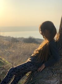 Side view of girl leaning on tree trunk while looking away outdoors