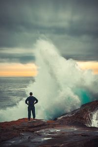 Rear view of man standing on rock while waves splashing against sky