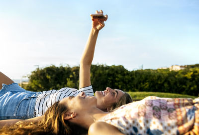 Smiling young woman taking selfie with female friend while relaxing on field during sunny day