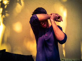 Young man covering eyes while standing against yellow wall with sunlight