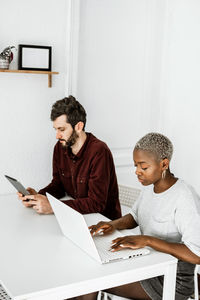 Side view from above of contemporary african american woman sitting at table and working with laptop with young man surfing digital table at home