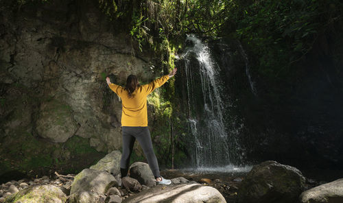 Woman standing next to a waterfall, with her arms open, in the middle of a forest during a sunny day