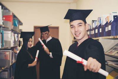 Portrait of smiling young man in graduation gown standing in library
