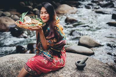 Smiling woman holding fresh food in bucket while sitting on rock amidst river