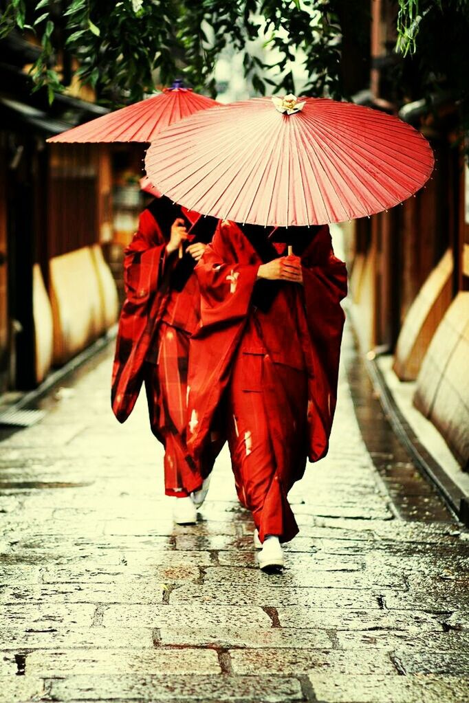 red, rear view, focus on foreground, street, full length, umbrella, the way forward, walking, building exterior, day, sidewalk, outdoors, footpath, built structure, cobblestone, incidental people, traditional clothing, sunlight, cultures