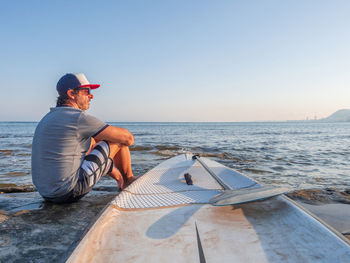 Side view of sporty mature man in sunglasses with big surfboard sitting in the seaside coast looking at the horizon in alicante spain