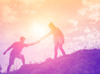 Man and woman with arms raised standing on land against sky during sunset