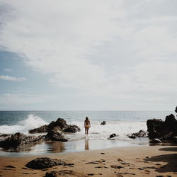 Rear view of young woman wearing bikini standing on shore at beach against sky