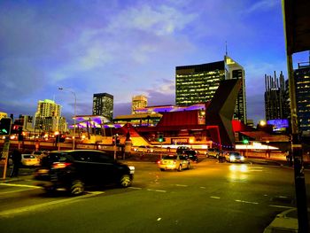 Cars on road by illuminated buildings against sky at dusk