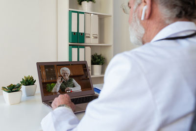 Back view of crop anonymous elderly male doctor in wireless earbud speaking with woman on video call on netbook in clinic