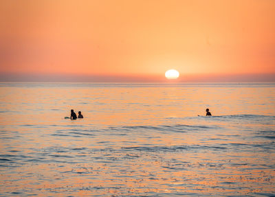 Silhouette people swimming in sea against orange sky