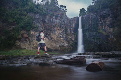 Full length of man with backpack looking at waterfall while standing on rock