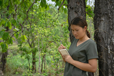 Woman writing in book while standing by tree trunk in forest