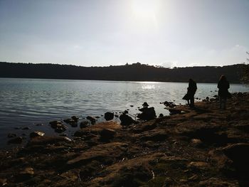 Silhouette people standing on rock by lake against sky during sunset