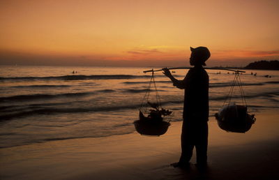 Silhouette boy with carrying pole standing on shore against sky during sunset