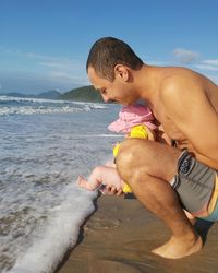 Full length of shirtless man with daughter crouching on beach against sky