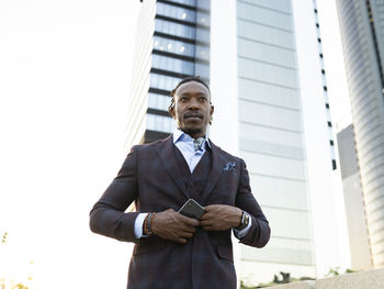 Low angle of serious african american male entrepreneur wearing classy suit standing with smartphone in hand on street in city center and looking away