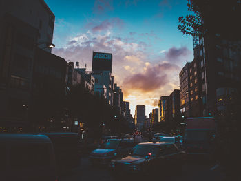 Traffic on city street by buildings against sky during sunset