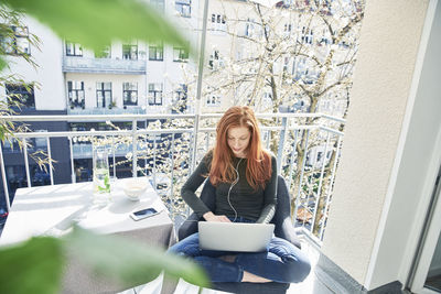 Redheaded woman sitting on balcony using laptop and earphones in spring