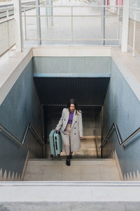From above of elegant stylish young female traveler with suitcase walking up stairs at train station