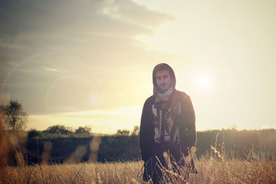 Portrait of young man standing amidst dried plants on field against sky during sunset