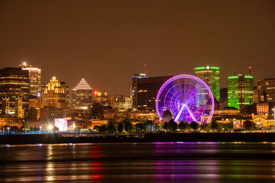 Illuminated ferris wheel by river against buildings at night