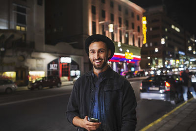 Portrait of happy man holding smart phone while standing on city street at night