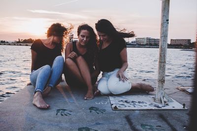 Young woman sitting on shore against sky during sunset