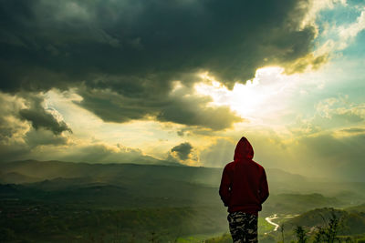 Rear view of young man standing on mountain against cloudy sky during sunset