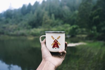 Close-up of hand holding mug over lake