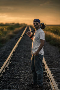 Portrait of young man with camera standing at railroad tracks against sky during sunset